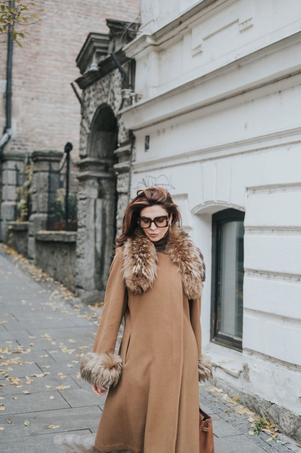 Lydia Esthetics Embassy A woman wearing a brown coat with fur trim and sunglasses walks on a sidewalk beside a white building, autumn leaves scattered on the ground—reminiscent of the elegance found at a luxury facial spa NYC locals adore. New York