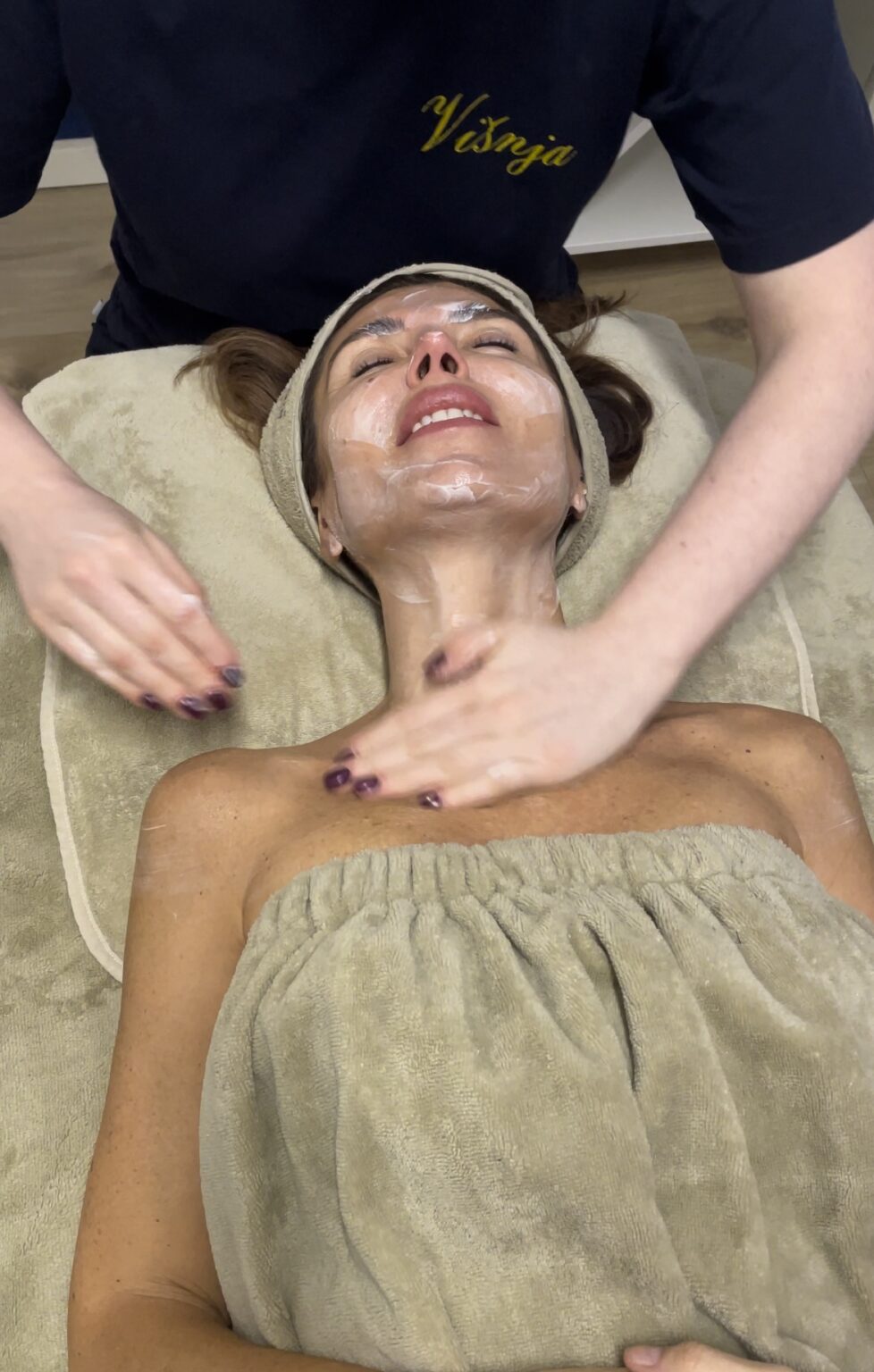 Lydia Esthetics Embassy A person lies on a spa table at a luxury facial spa NYC with cream applied to their face and neck while a practitioner massages their neck and shoulders. New York