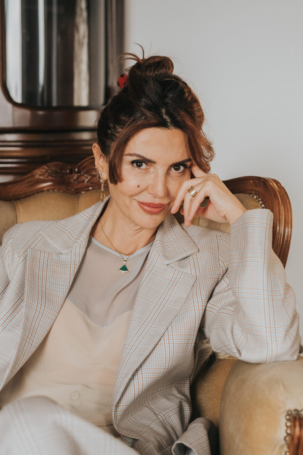Lydia Esthetics Embassy A woman with brown hair in a checked suit sits on an armchair, resting her head on her hand and looking at the camera with a slight smile—like she's just returned from a luxury facial spa NYC experience. New York