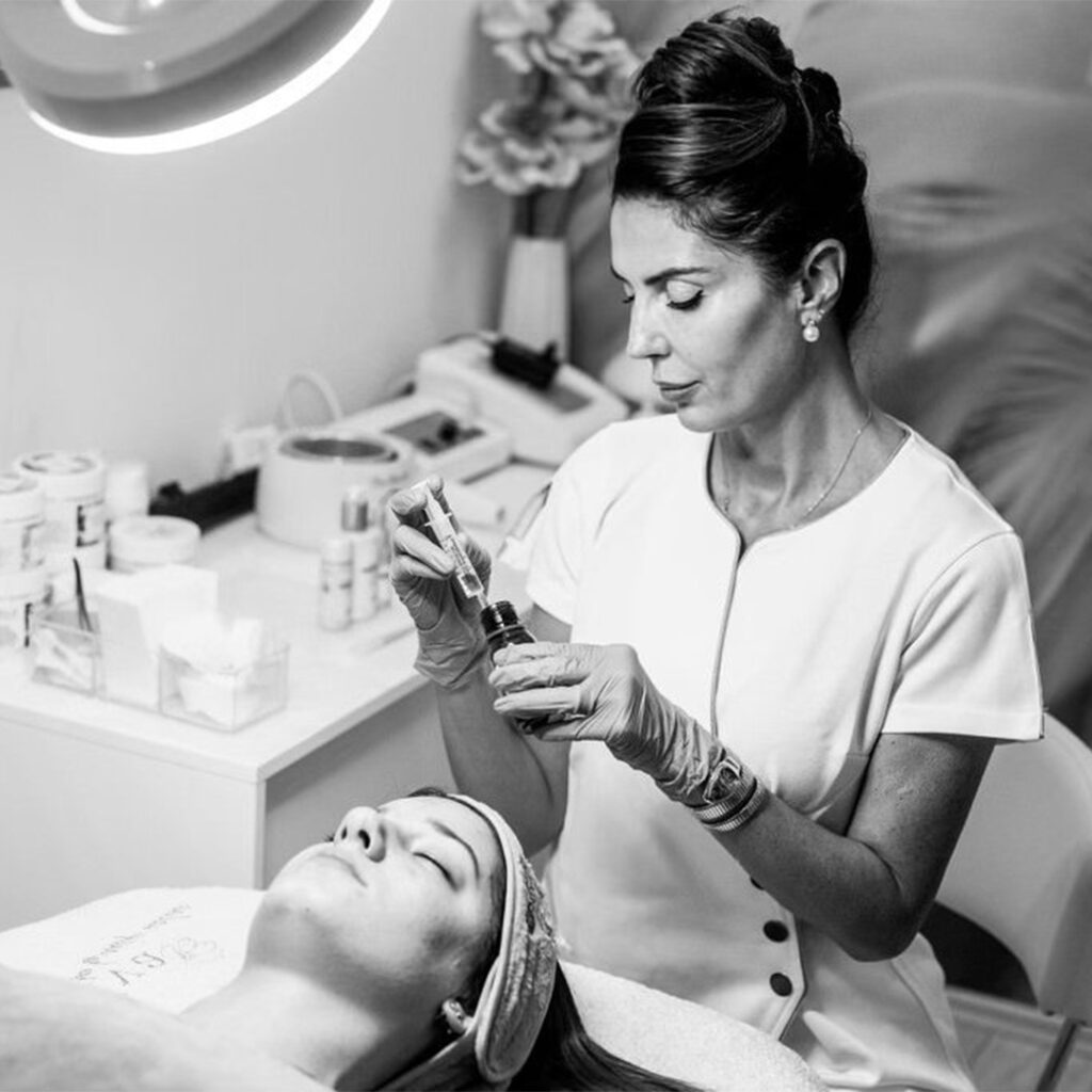 Lydia Esthetics Embassy A woman in medical attire prepares an injection near a reclining patient’s face, illustrating advanced treatments like microneedling with exosomes in New York within a modern clinical setting. New York
