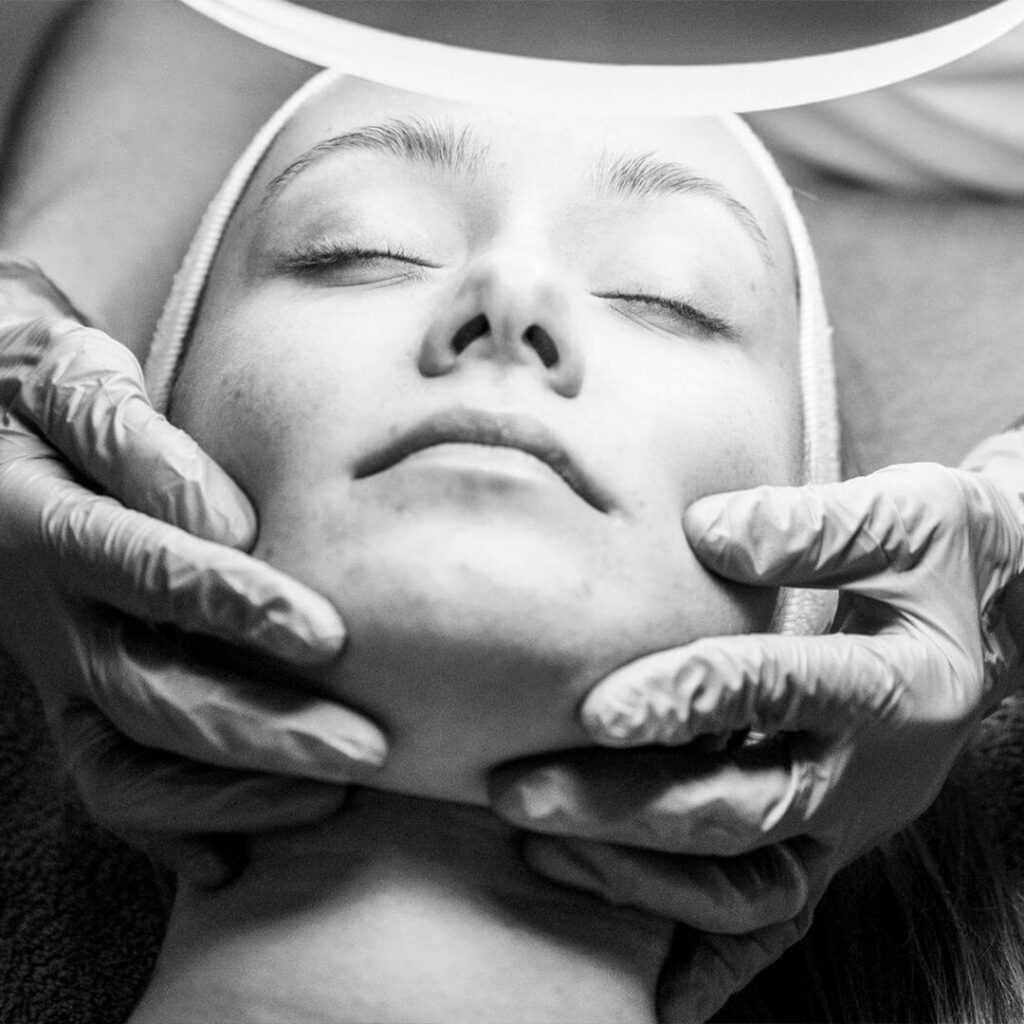 Lydia Esthetics Embassy A person with closed eyes receives a facial treatment, as gloved hands gently hold their face under a bright examination light at Esthetics Embassy New York. New York