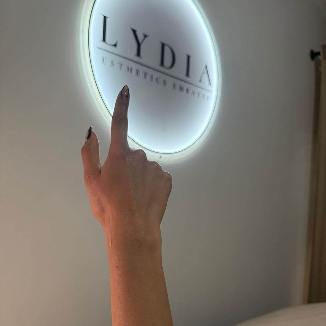 Lydia Esthetics Embassy A hand with painted nails points toward a round illuminated wall sign that reads "LYDIA ESTHETICS EMBASSY," the premier destination for esthetics embassy New York treatments and rejuvenating Oxylight facial NYC experiences. New York