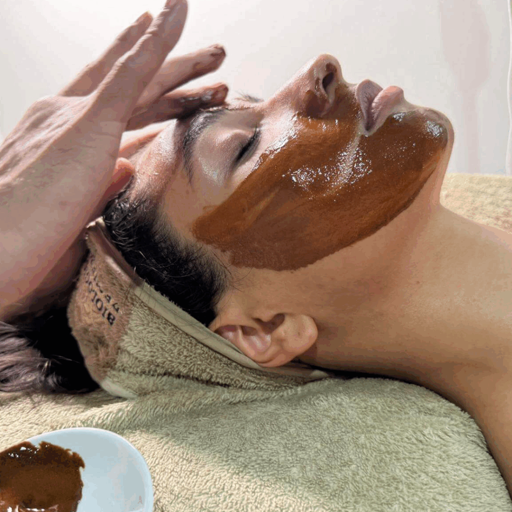 Lydia Esthetics Embassy A person lying on a towel with eyes closed receives a luxurious facial treatment at a luxury facial spa NYC as a dark brown mask is applied to their face. New York
