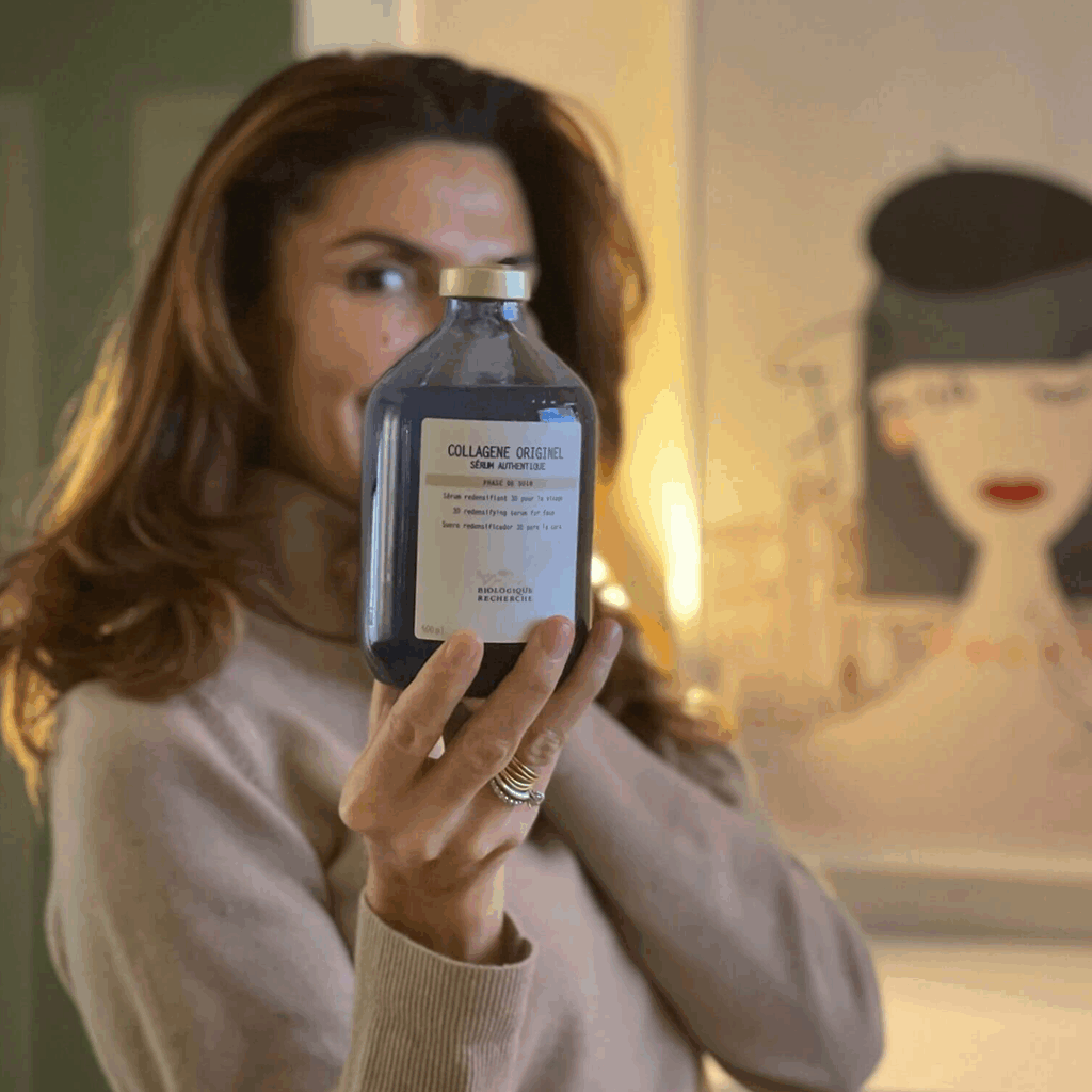 Lydia Esthetics Embassy A person in a beige sweater holds up a bottle labeled "Collagene Originel," suggesting ties to luxury facial spa NYC treatments, with their face blurred and an art piece of a woman visible in the background. New York