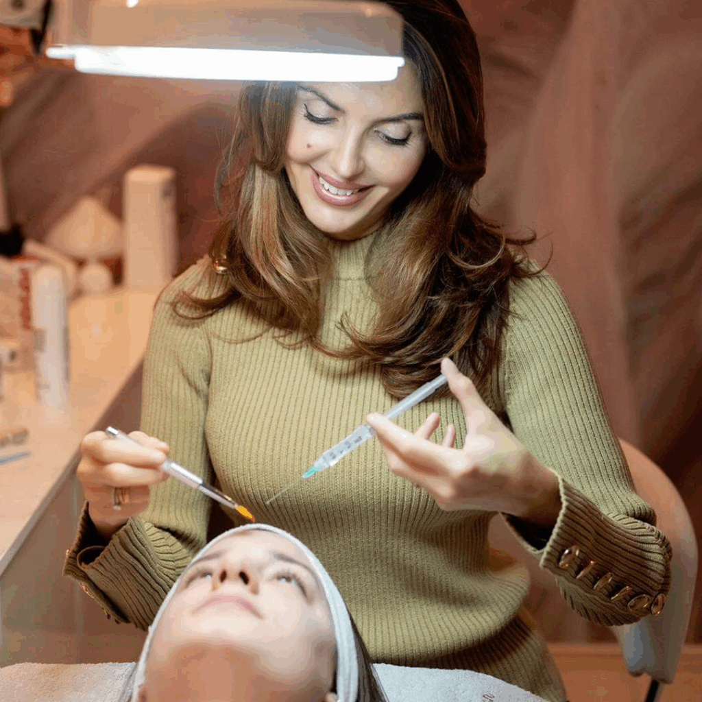 Lydia Esthetics Embassy A woman in a tan sweater prepares to apply a facial treatment with a brush and syringe under a bright lamp at Esthetics Embassy New York, known for its oxylight facial NYC and microcurrent facial non surgical facelift. New York