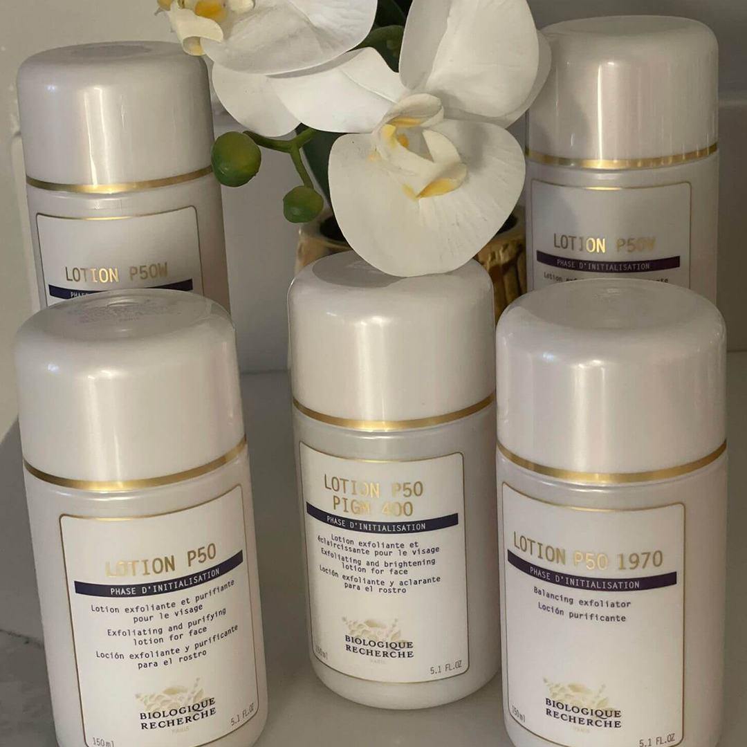 Lydia Esthetics Embassy Six bottles of Biologique Recherche skincare lotions, including variations of Lotion P50, are arranged on a shelf with a white orchid in the background at Esthetics Embassy New York. New York