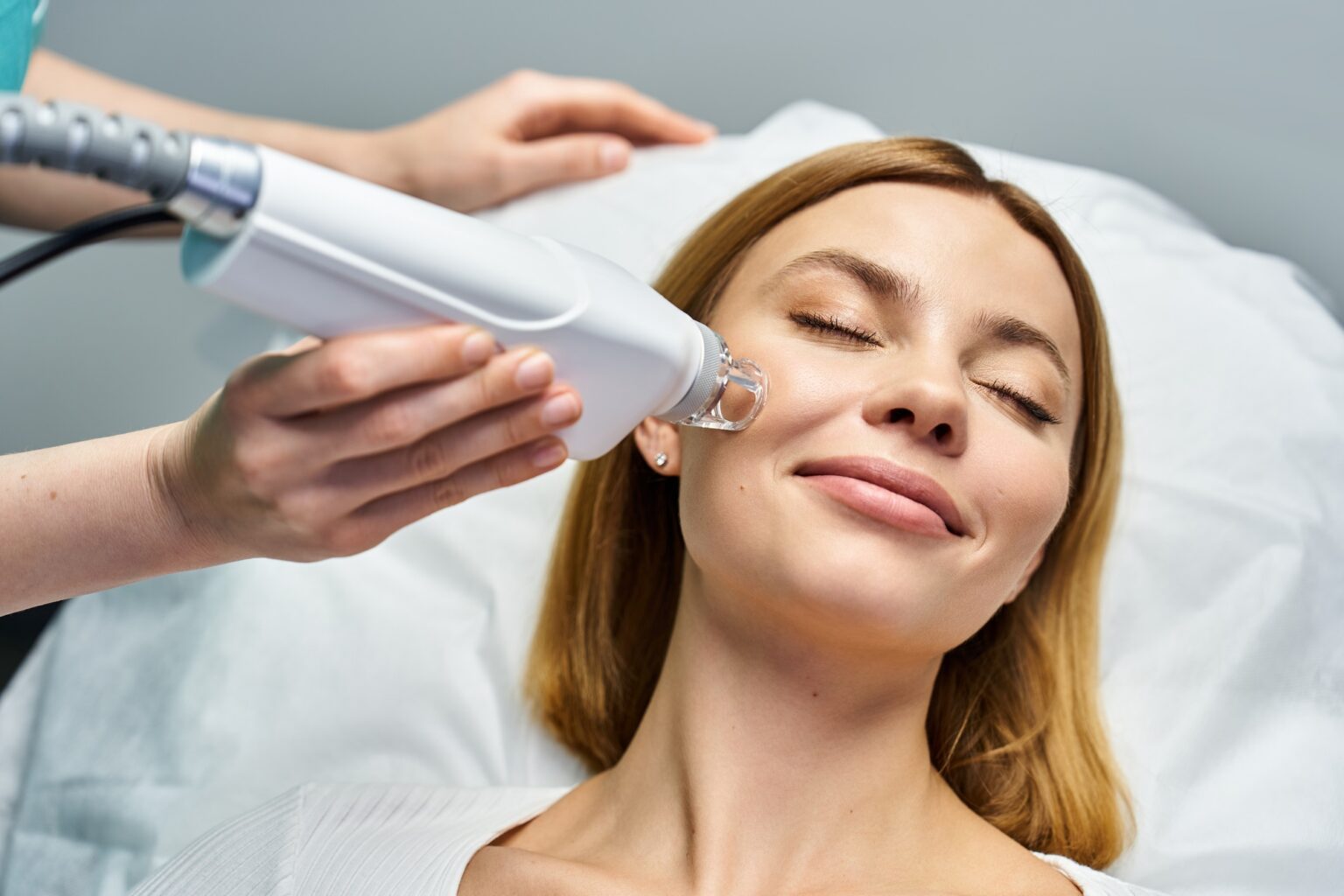 Lydia Esthetics Embassy A woman lies on a clinic bed with her eyes closed while a practitioner uses a handheld device on her cheek during an Oxylight facial NYC treatment. New York
