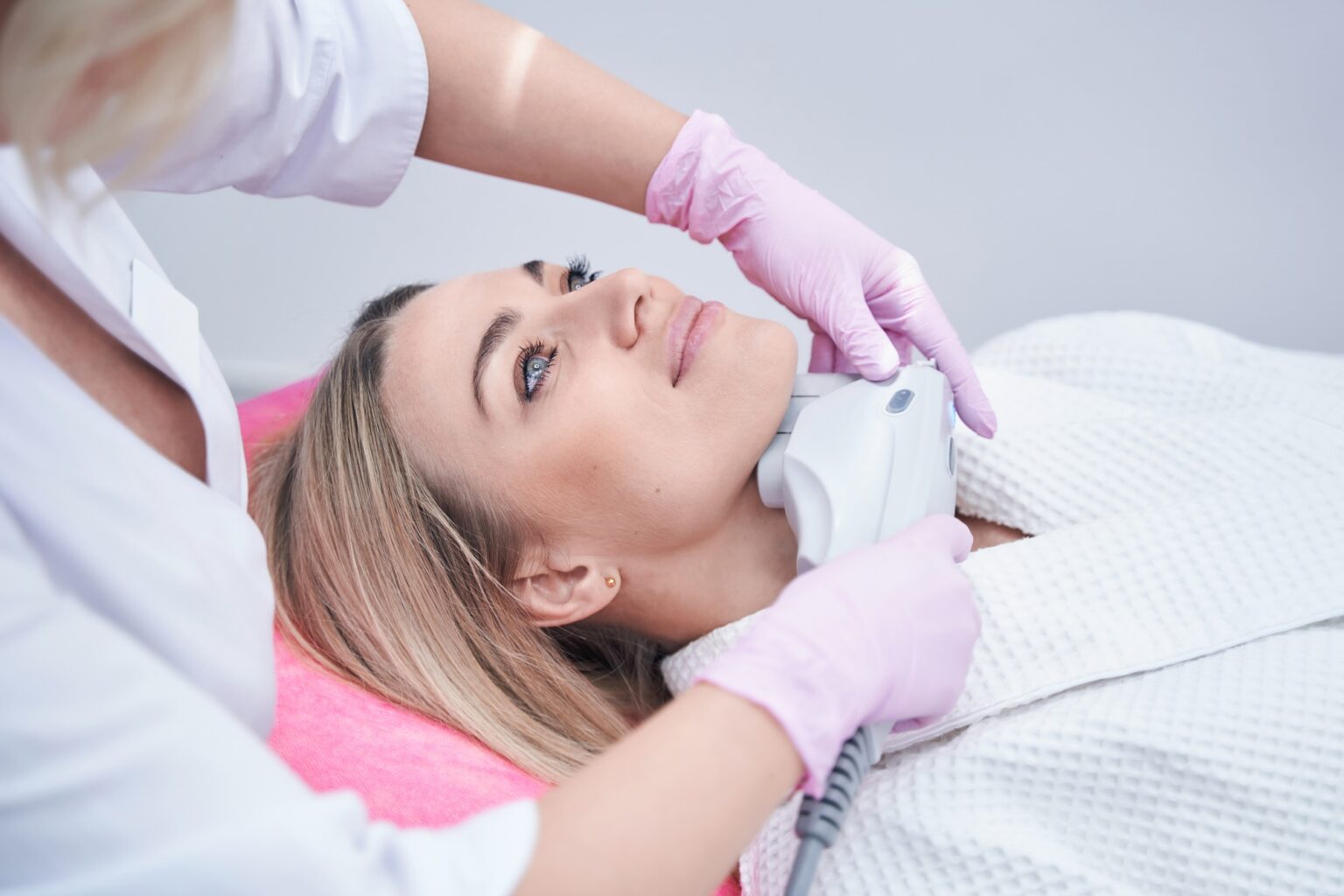 Lydia Esthetics Embassy A woman in a white robe receives an Oxylight facial NYC treatment with a handheld device applied to her chin by a technician wearing pink gloves. New York