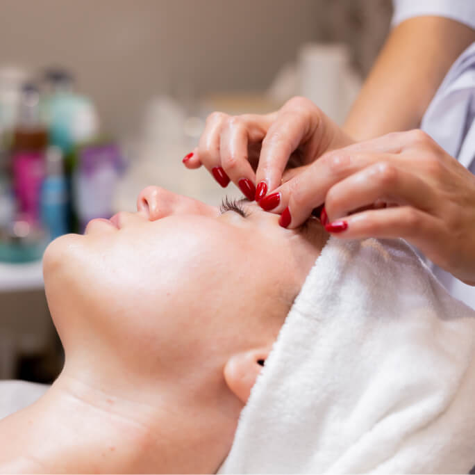 Lydia Esthetics Embassy A person lies with eyes closed and a towel wrapped around their head at Esthetics Embassy New York while another person applies eyelash extensions. New York