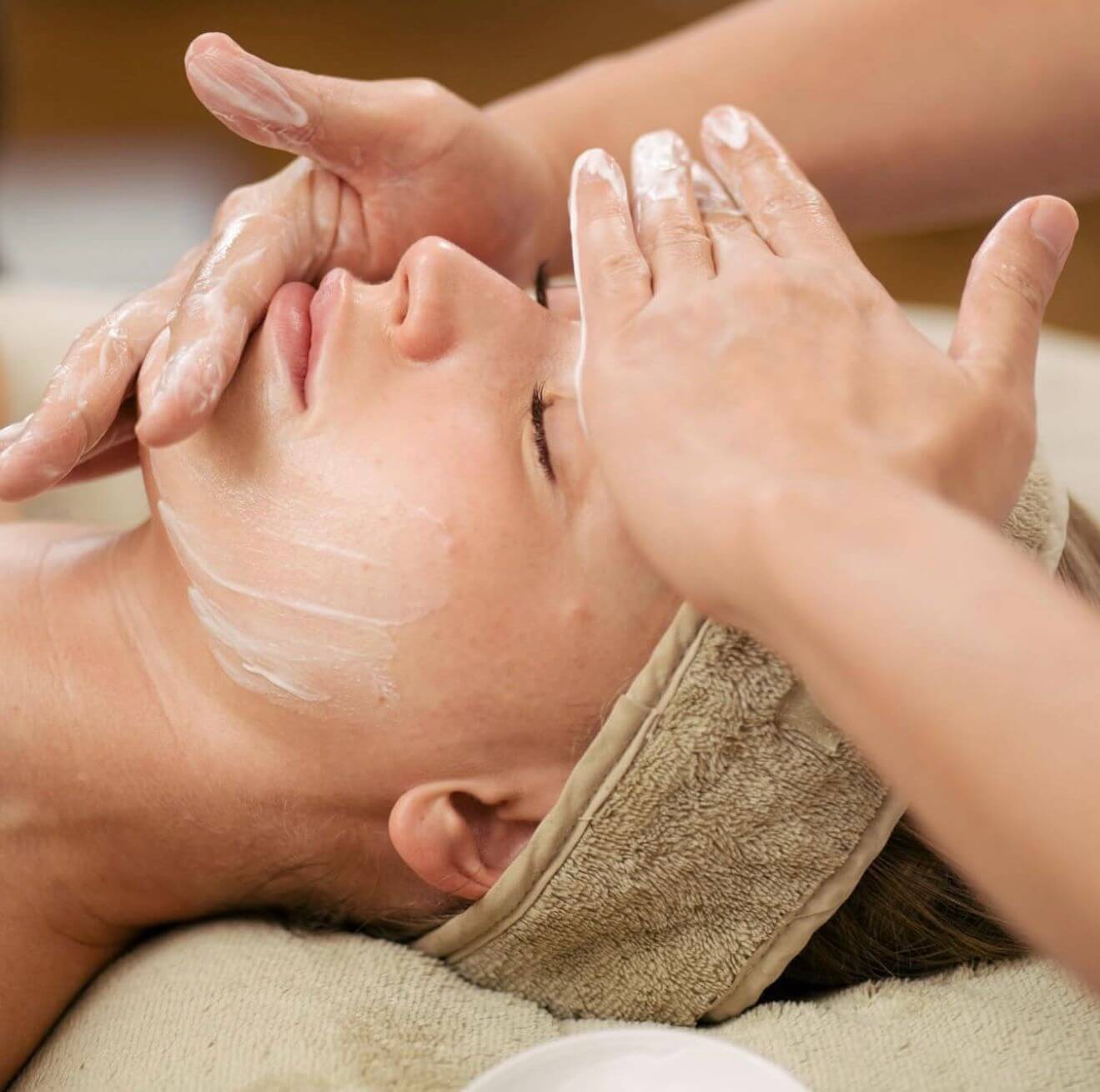 Lydia Esthetics Embassy Person receiving a facial treatment, with cream gently massaged into their skin by another’s hands. The individual, eyes closed and wearing a beige headband, enjoys the soothing ambiance of a luxury facial spa NYC. New York