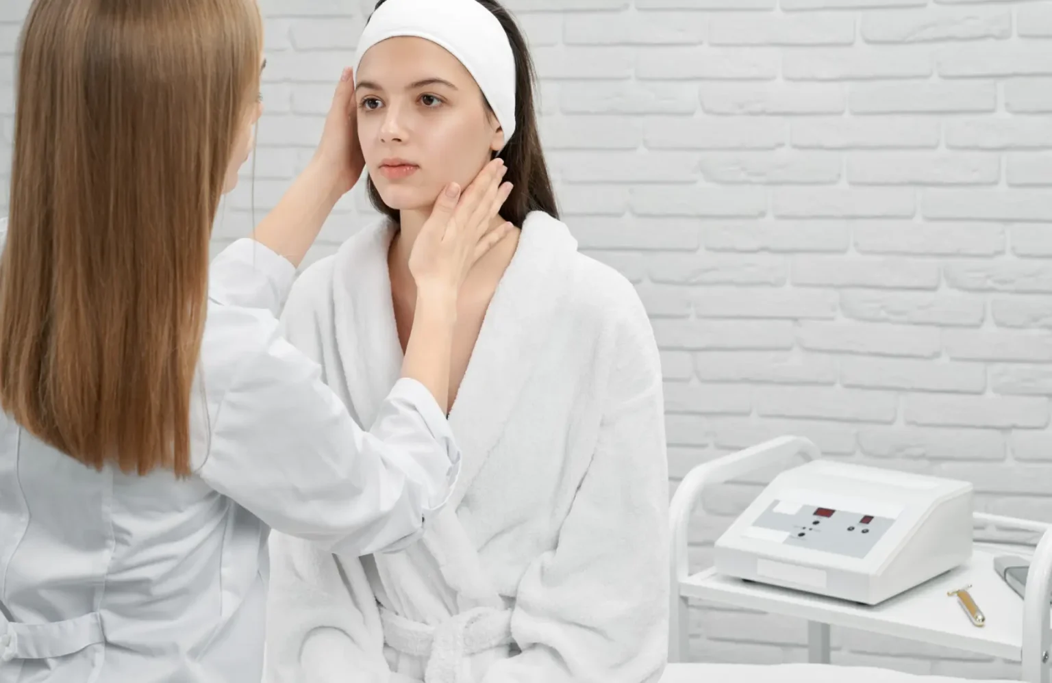 Lydia Esthetics Embassy A woman in a white robe and headband is examined by a healthcare professional in a clinical setting, with medical equipment visible in the background. New York