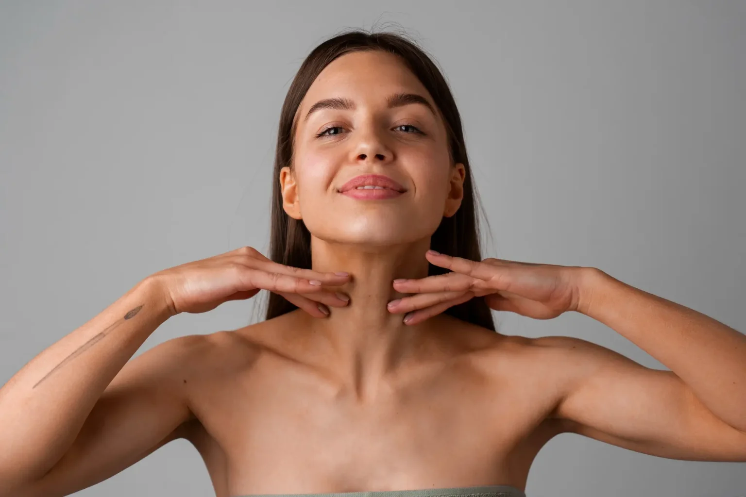 Lydia Esthetics Embassy A woman with long brown hair touches her neck with both hands, looking forward and smiling softly against a plain gray background. New York