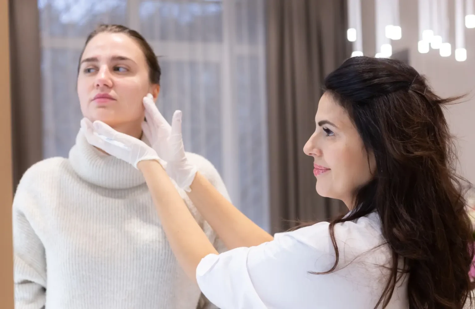 Lydia Esthetics Embassy A woman wearing gloves examines another woman's jawline and face in a bright indoor setting. New York