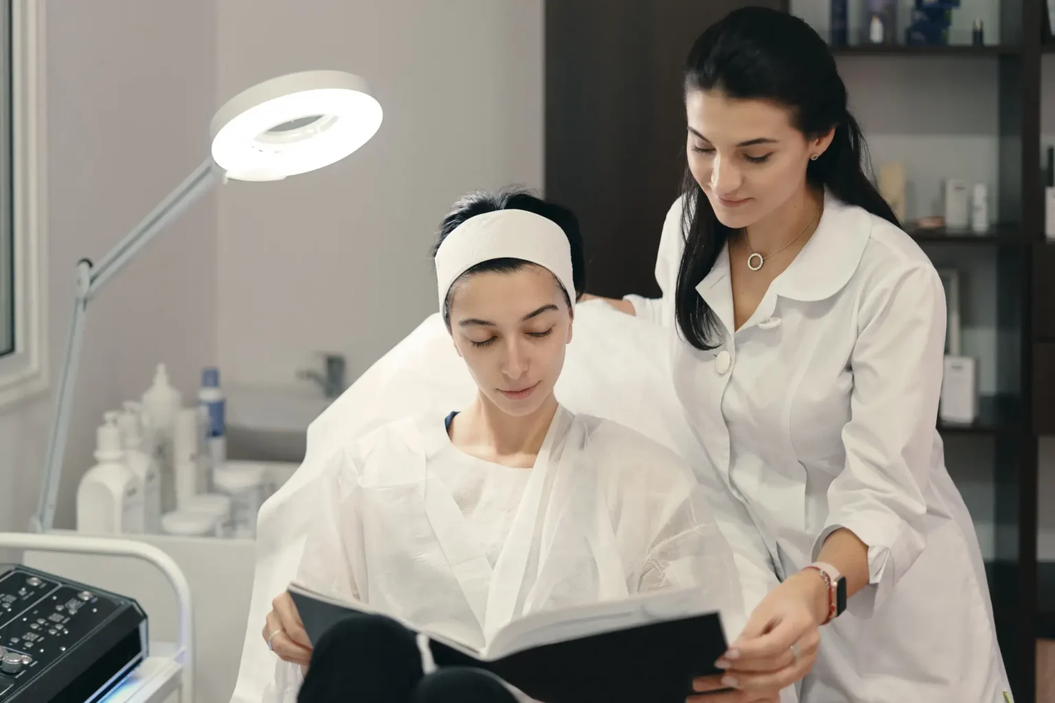 Lydia Esthetics Embassy Two women in a clinic, one sitting in a chair wearing a headband and gown reading a booklet, while the other, in a white coat, stands beside her, offering assistance. New York