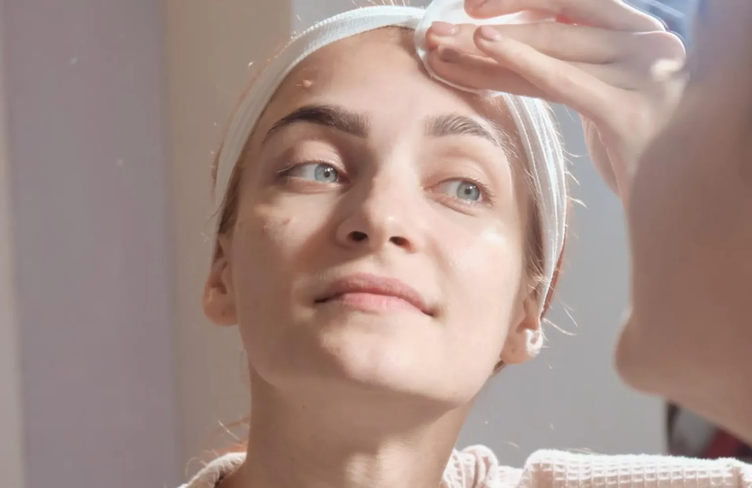 Lydia Esthetics Embassy A woman with a headband applies a cotton pad to her forehead while looking in the mirror. New York