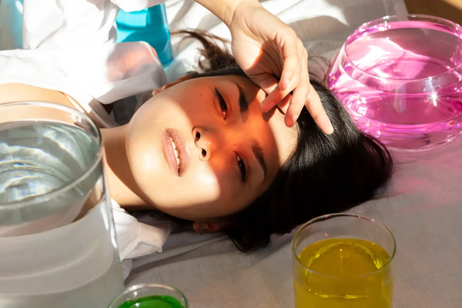 Lydia Esthetics Embassy A woman lies on a surface with her hand on her forehead, surrounded by glass bowls containing green, yellow, and pink liquids. Sunlight casts shadows across her face. New York