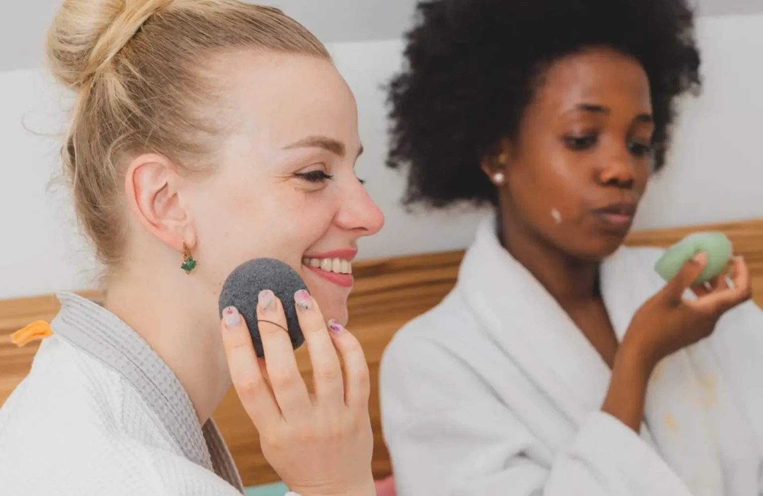 Lydia Esthetics Embassy Two women in bathrobes apply skincare products; one uses a facial sponge while the other holds a bar of soap. New York