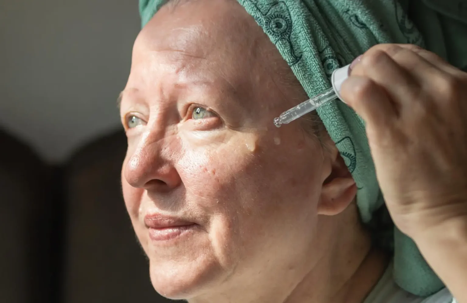 Lydia Esthetics Embassy An older woman with a green towel wrapped around her head applies facial serum under her eye using a dropper. New York