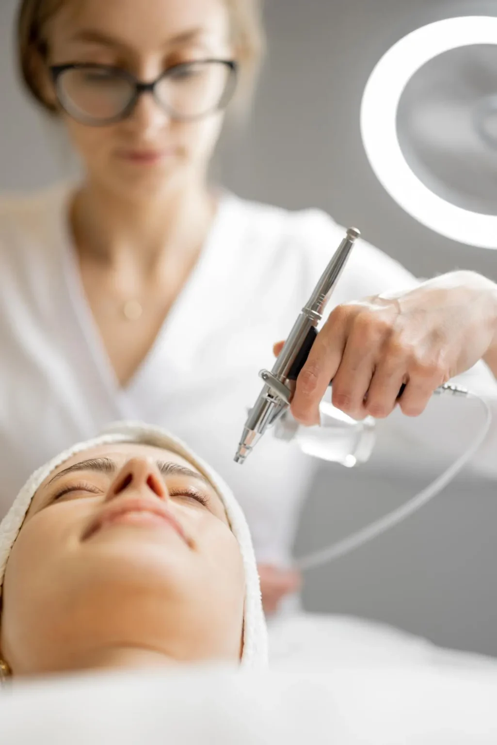 Lydia Esthetics Embassy A cosmetologist in glasses performs a facial procedure using a handheld device on a client lying down with closed eyes and a towel wrapped around her head. New York