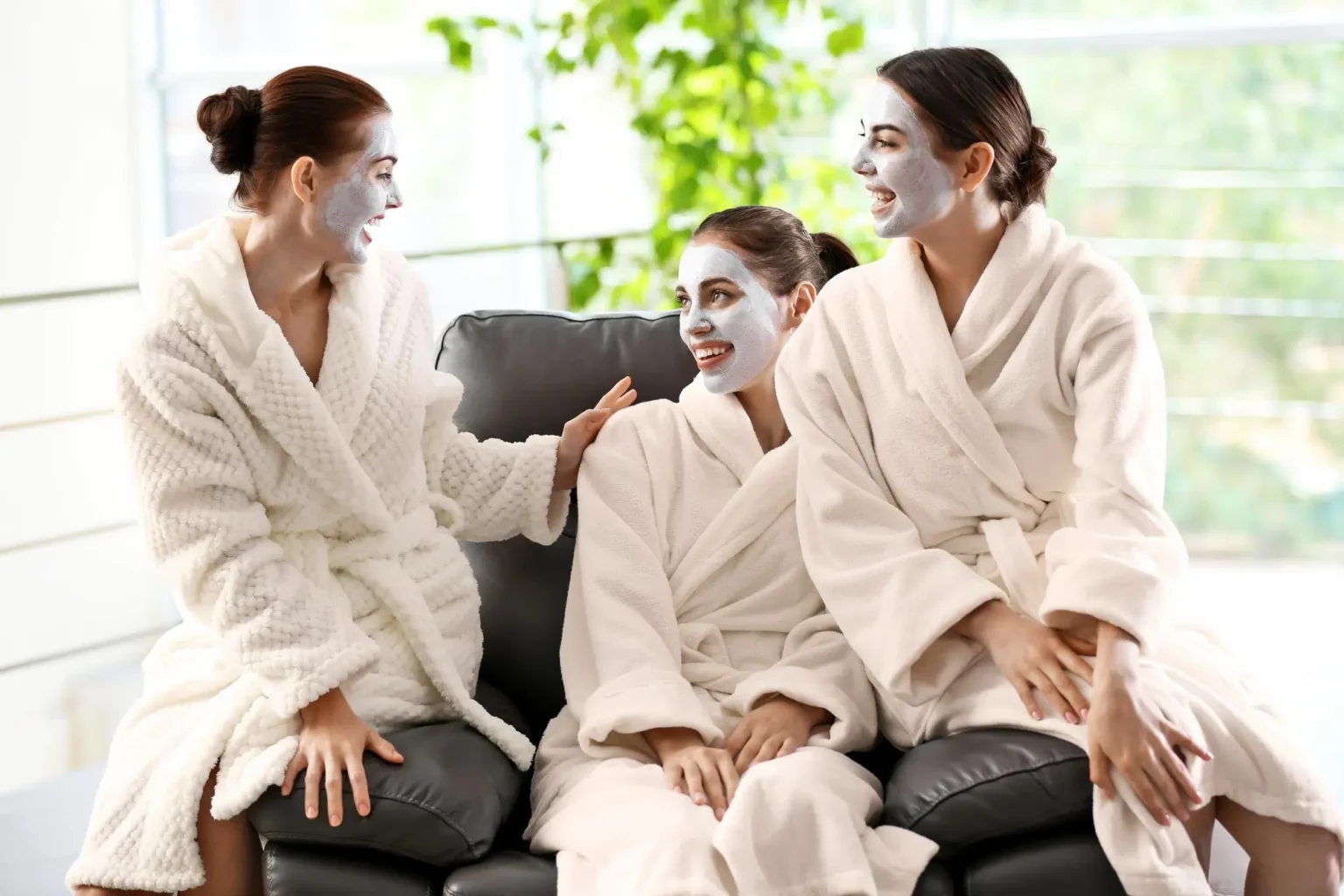 Lydia Esthetics Embassy Three people in white bathrobes with facial masks are sitting and chatting on a black couch in a bright room with green plants in the background. New York