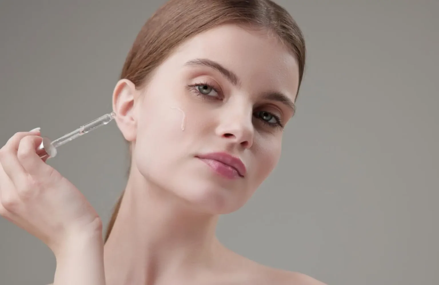 Lydia Esthetics Embassy A woman applies a skincare serum to her face using a glass dropper against a plain, neutral background. New York
