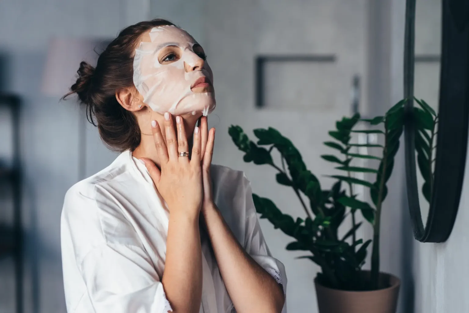 Lydia Esthetics Embassy A woman wearing a sheet mask and a white robe touches her neck while looking at herself in a mirror, with a potted plant in the background. New York