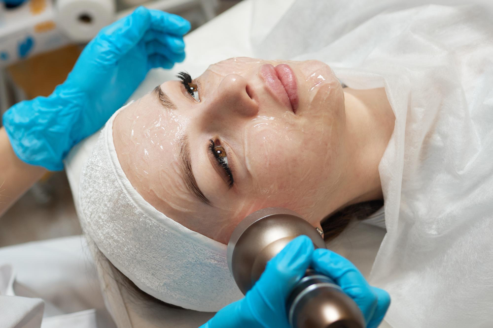 Lydia Esthetics Embassy A person lies on a treatment table with a clear facial mask, wearing a headband, while a gloved technician uses a handheld device on their face. New York