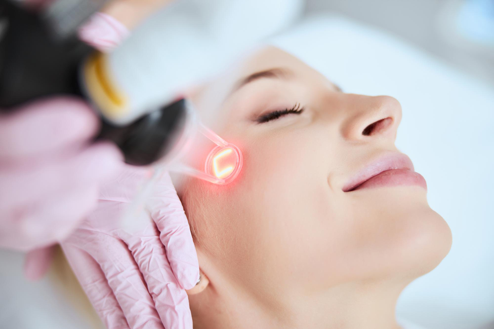 Lydia Esthetics Embassy A person receiving a laser skin treatment on their face by a gloved professional, with a red light focused on the cheekbone area. New York