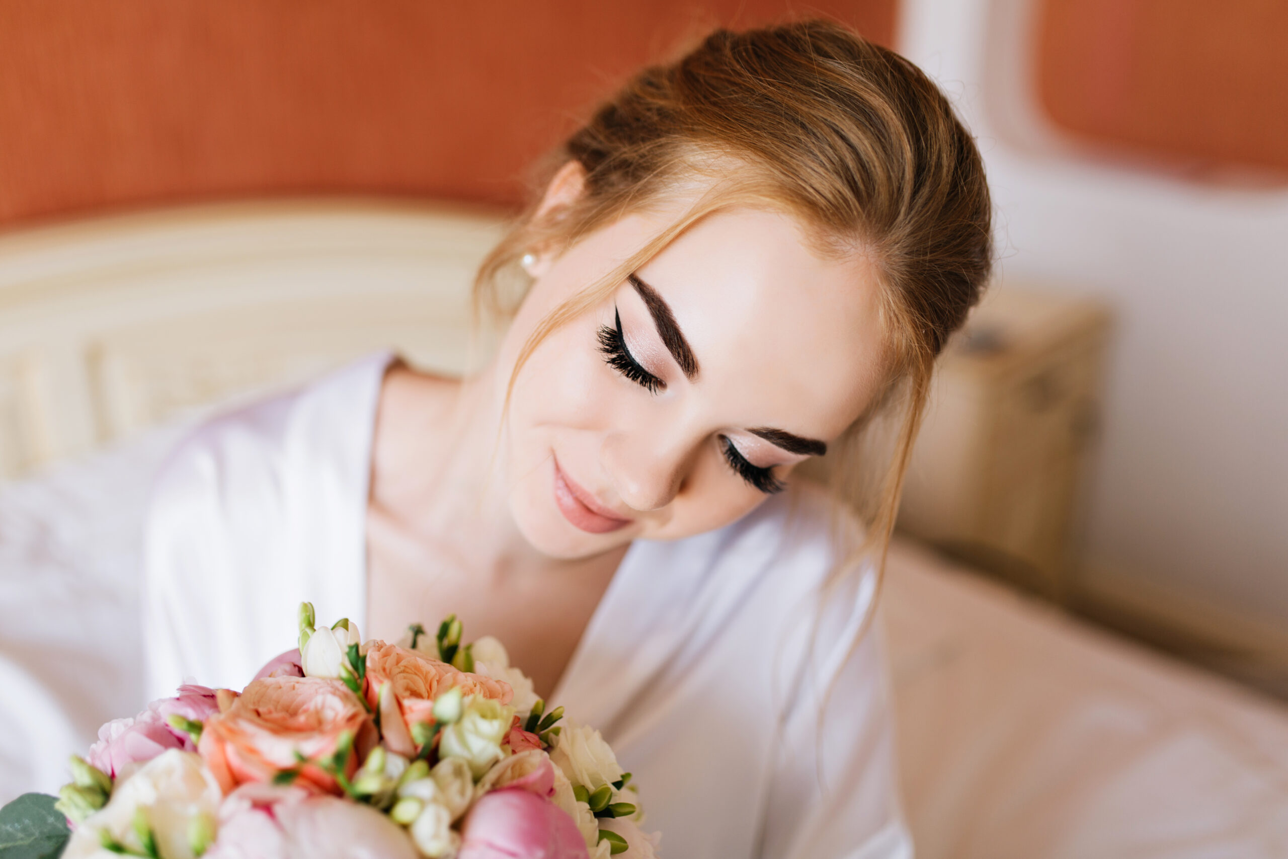 Lydia Esthetics Embassy A woman with closed eyes and light makeup sits on a bed, holding a bouquet of pink and white flowers, wearing a white robe. New York