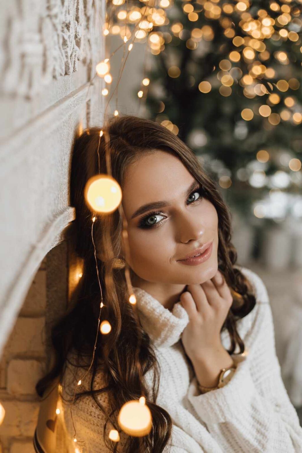 Lydia Esthetics Embassy Woman in a white sweater leans against a fireplace, surrounded by string lights, with a blurred background of holiday lights and decorations. New York