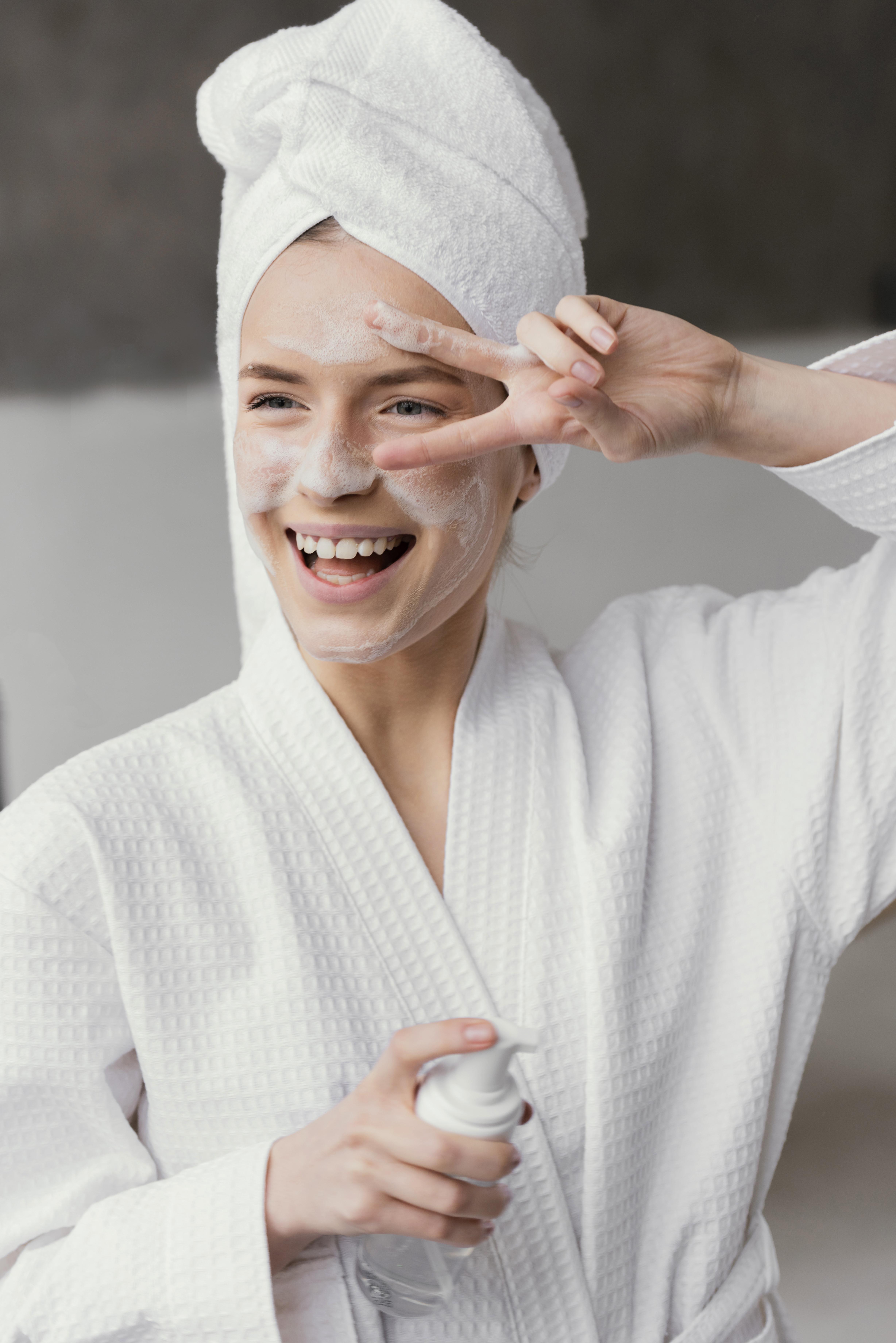 Lydia Esthetics Embassy A person in a white bathrobe and towel on their head applies cream to their face, smiling and making a peace sign with their hand. New York