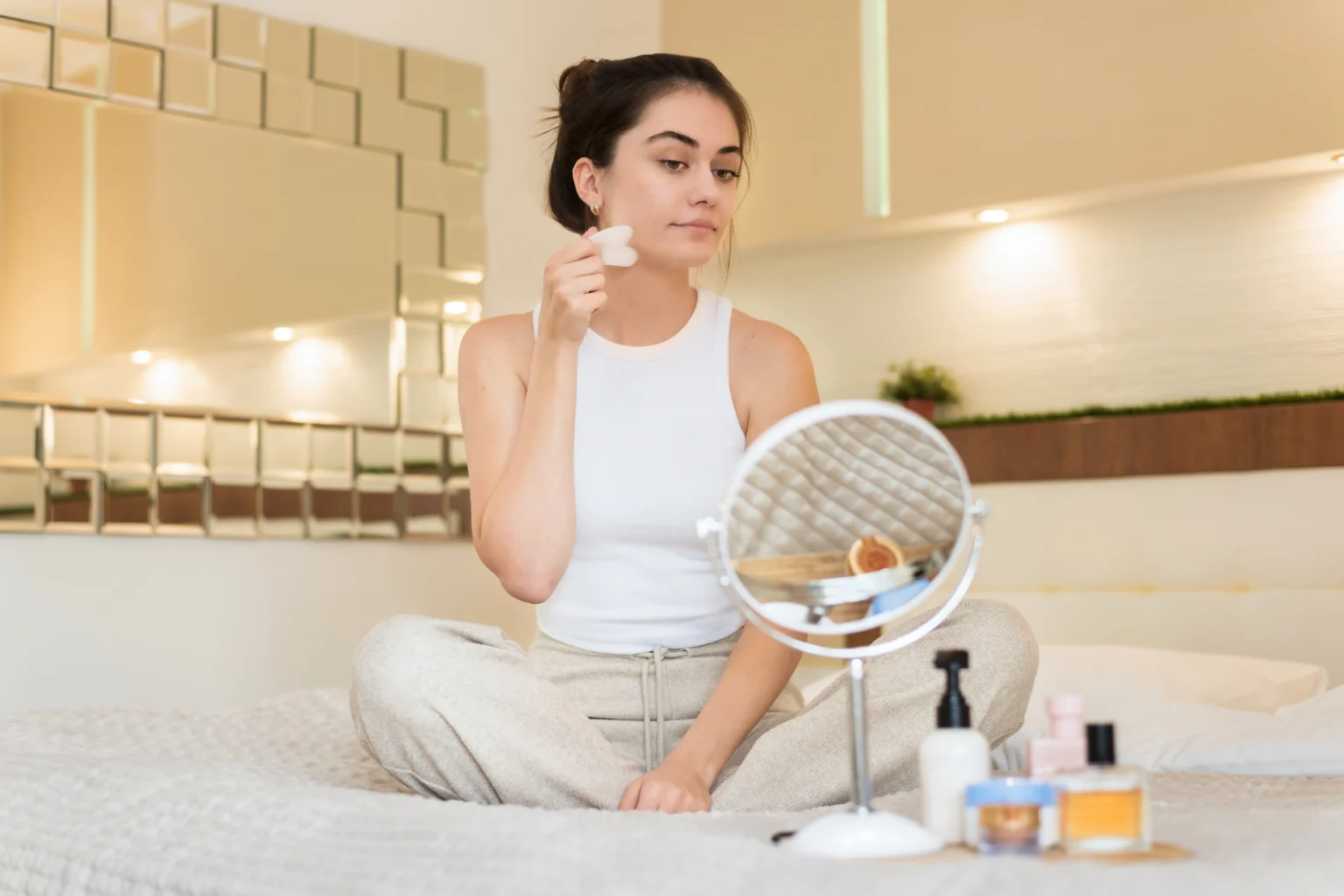 Lydia Esthetics Embassy A woman in casual clothes sits on a bed, using a facial roller in front of a round mirror, with various skincare products on the blanket. New York