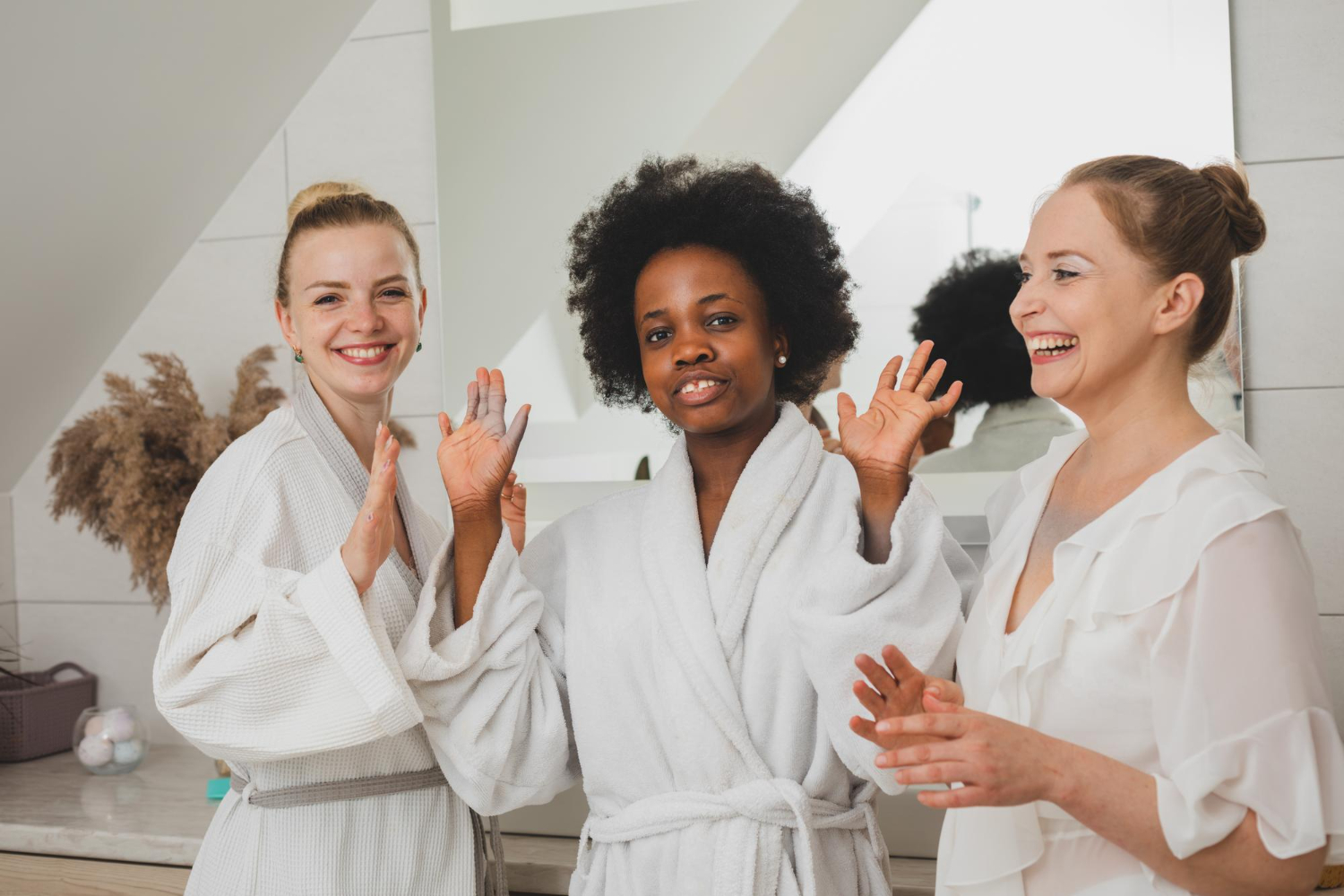 Lydia Esthetics Embassy Three women in bathrobes stand in a bathroom, smiling and posing together in front of a mirror. New York