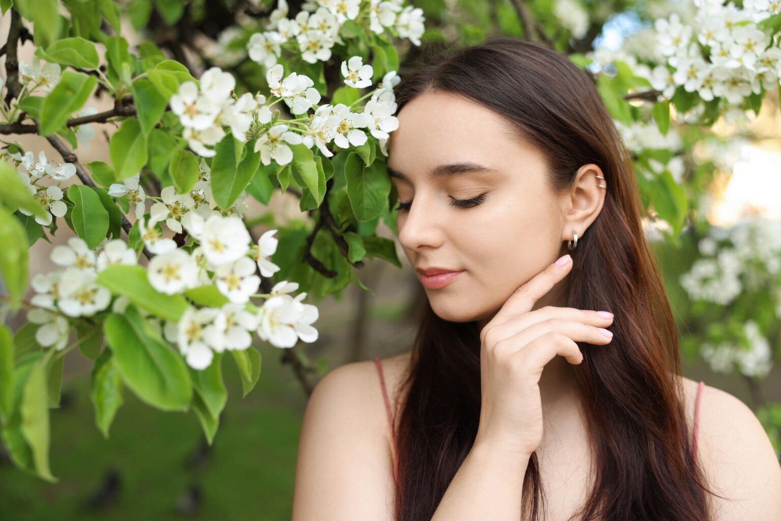 Lydia Esthetics Embassy A woman with long brown hair stands near a blossoming tree, eyes closed, with her hand gently touching her chin. White flowers and green leaves surround her. New York
