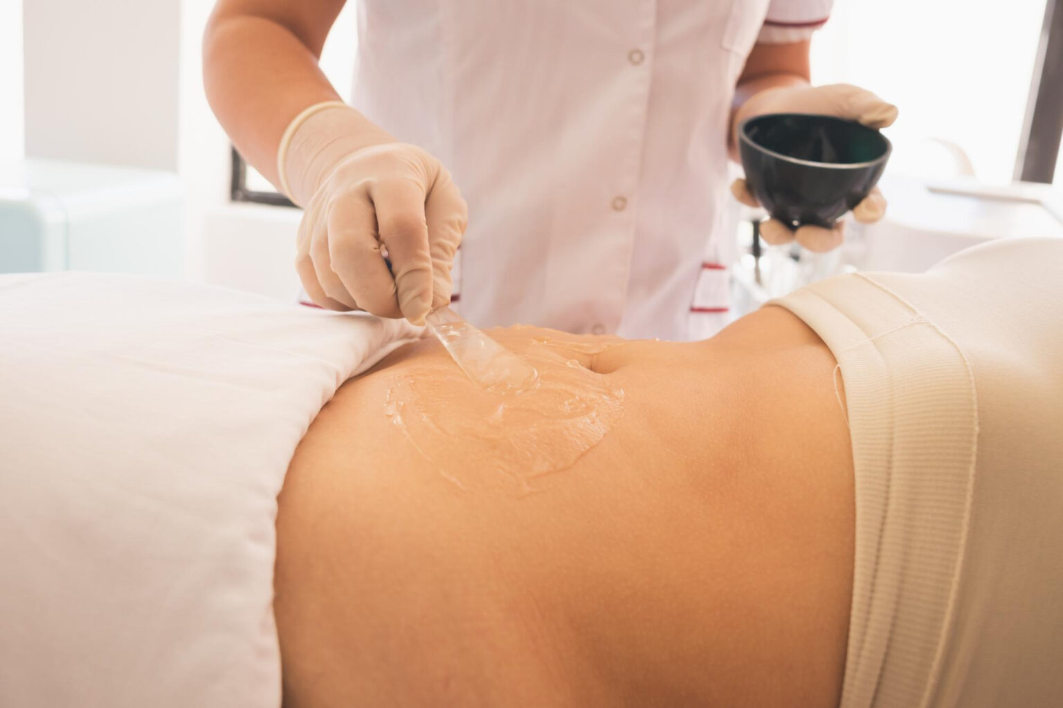 Lydia Esthetics Embassy A person in gloves applies a clear gel to someone's lower back using a spatula in a clinical or spa setting. New York