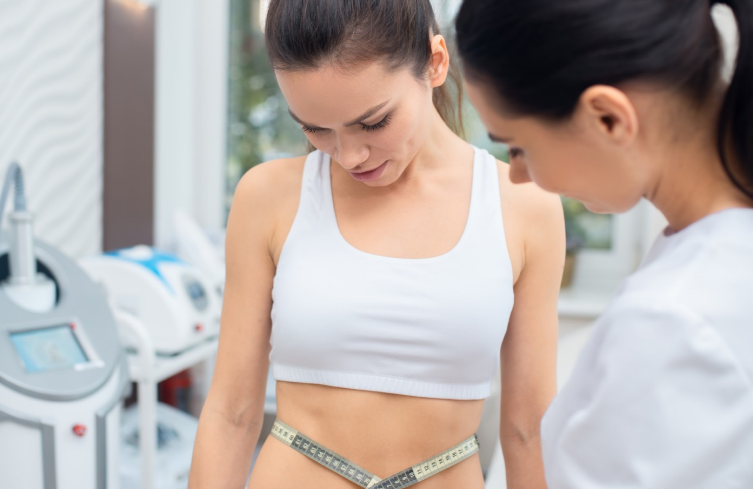 Lydia Esthetics Embassy Two women in a clinic setting measure one woman's waist with a tape measure. The woman being measured is wearing a white sports bra. New York