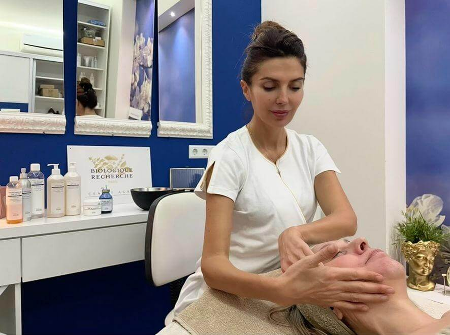 Lydia Esthetics Embassy A woman in a white uniform performs a facial massage on a client in a spa treatment room with skincare products on the counter. New York