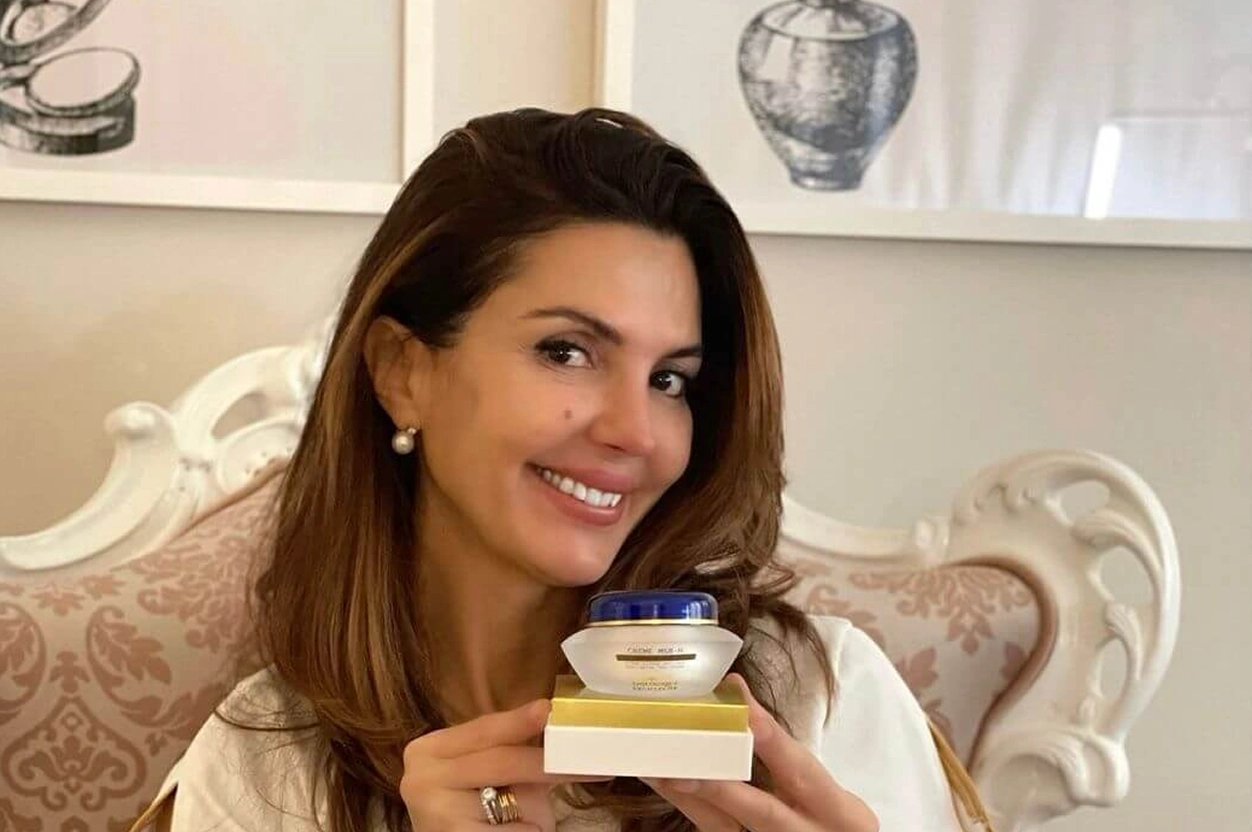 Lydia Esthetics Embassy Woman sitting on a patterned sofa, smiling and holding up a skincare cream jar with both hands. Two framed artworks hang on the wall behind her. New York