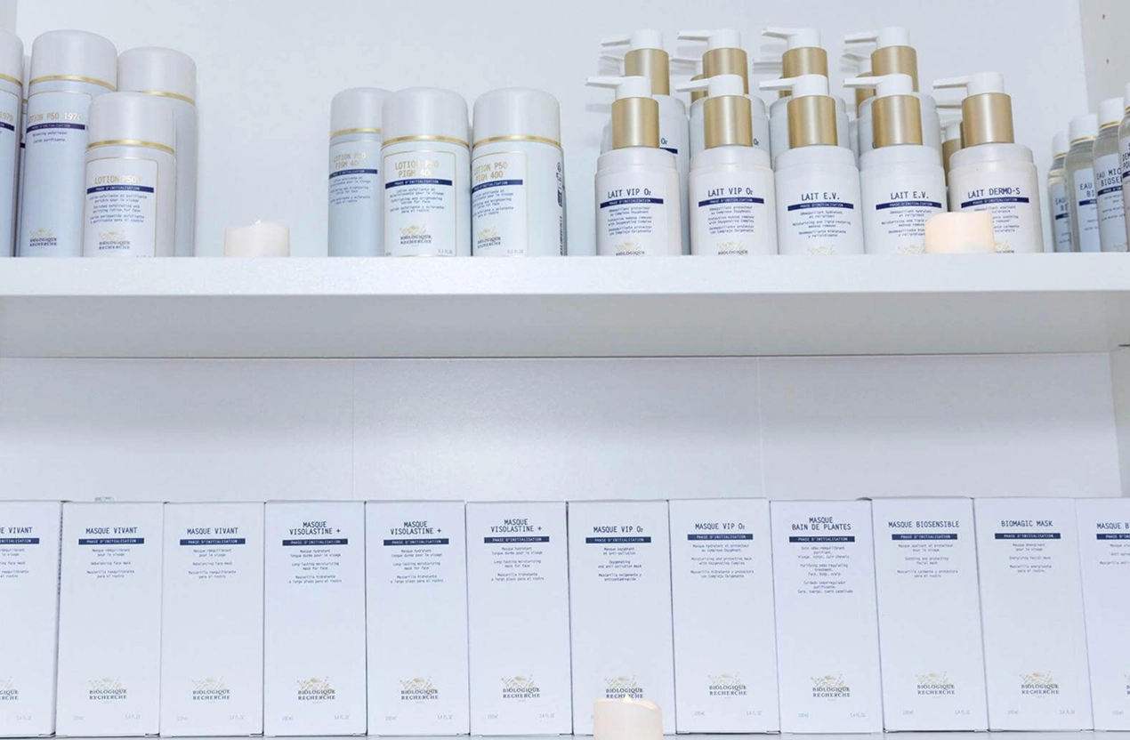 Lydia Esthetics Embassy Two shelves display neatly arranged bottles and boxes of skincare products with white and gold packaging. New York