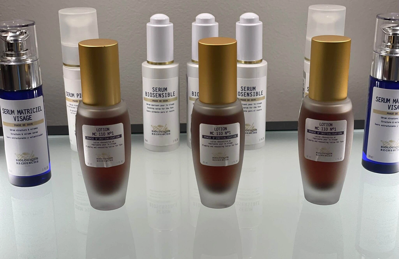 Lydia Esthetics Embassy Several bottles of skincare products, including three labeled "Lotion MC 110 N°1" with gold caps, are arranged on a glass shelf against a neutral background. New York