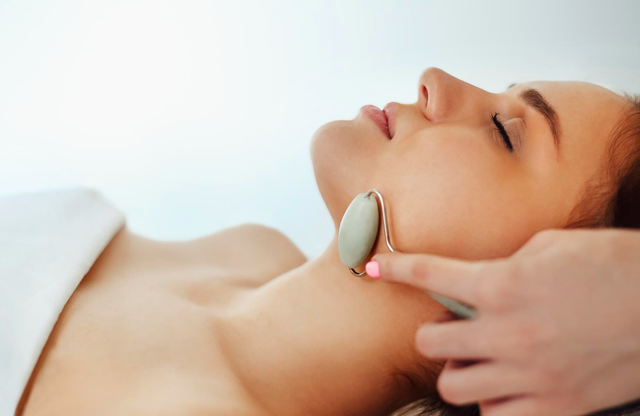 Lydia Esthetics Embassy A woman with closed eyes lies down while another person uses a jade roller on her jawline and neck. New York