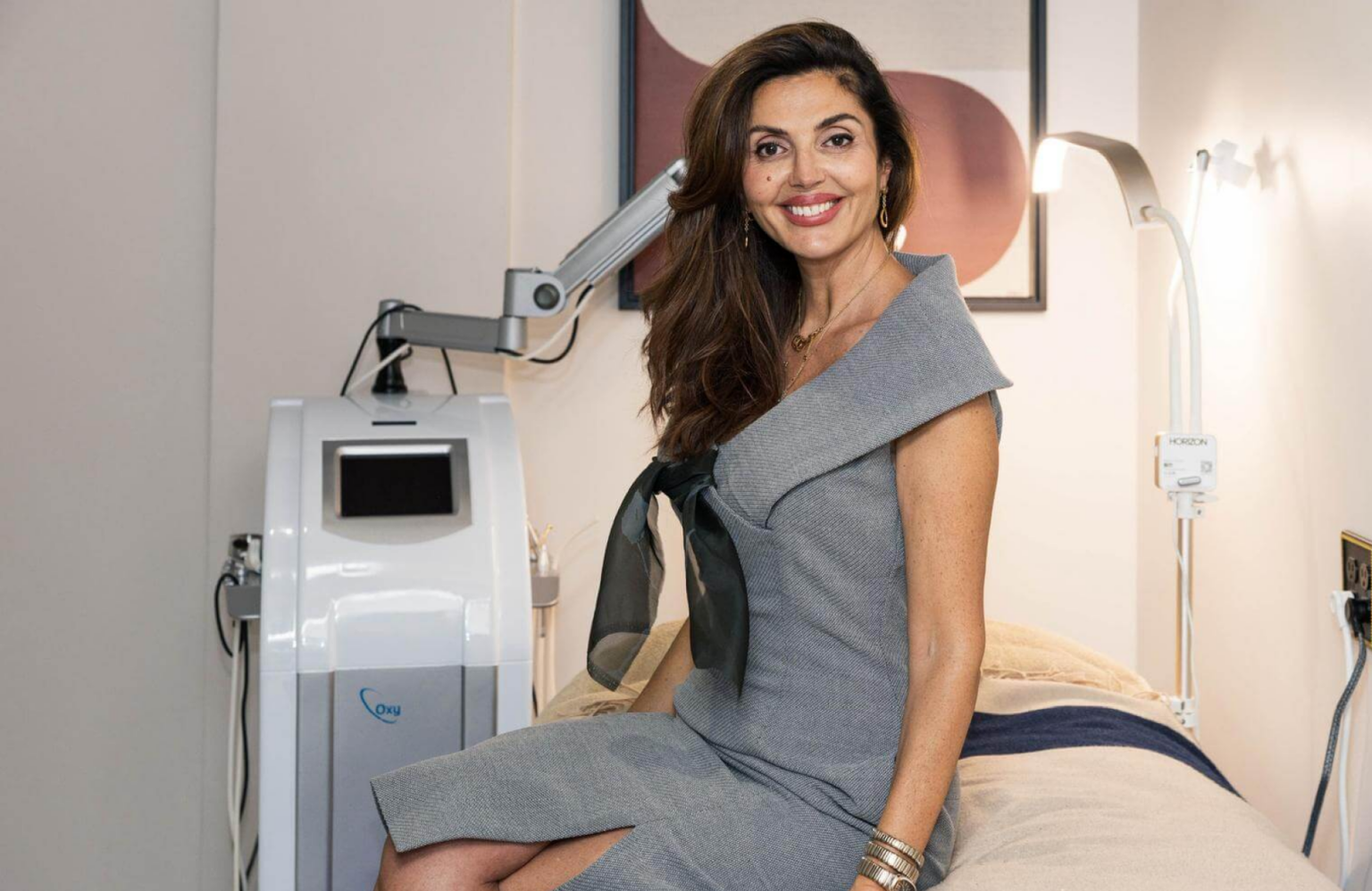 Lydia Esthetics Embassy Woman in a gray dress sits on a medical treatment bed next to skin resurfacing equipment in a modern clinic room. New York