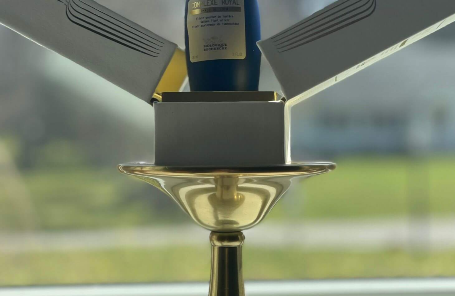 Lydia Esthetics Embassy A blue bottle of lactic acid cleanser in an open white box sits on top of an upside-down gold candlestick in front of a window with a blurred outdoor background. New York