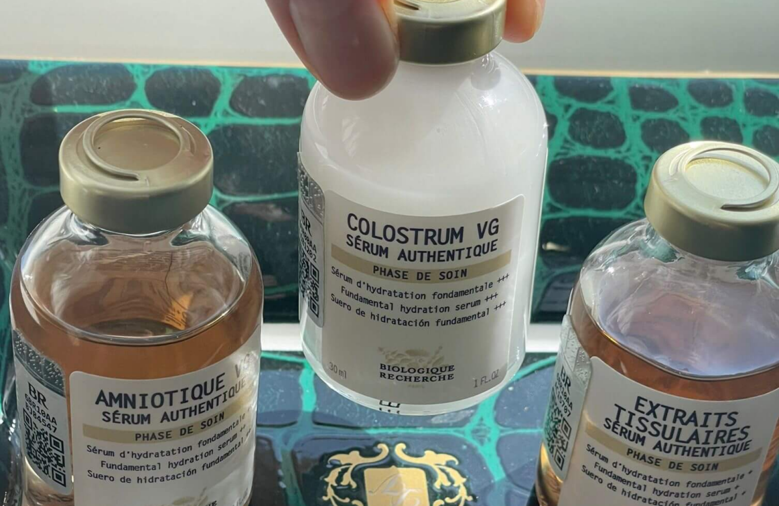 Lydia Esthetics Embassy Three glass bottles labeled “AMNIOTIQUE,” “COLOSTRUM VG,” and “EXTRAITS TISSULAIRES” are held on a decorative surface, containing skincare serums by Biologique Recherche, each formulated with skin barrier repair ingredients. New York