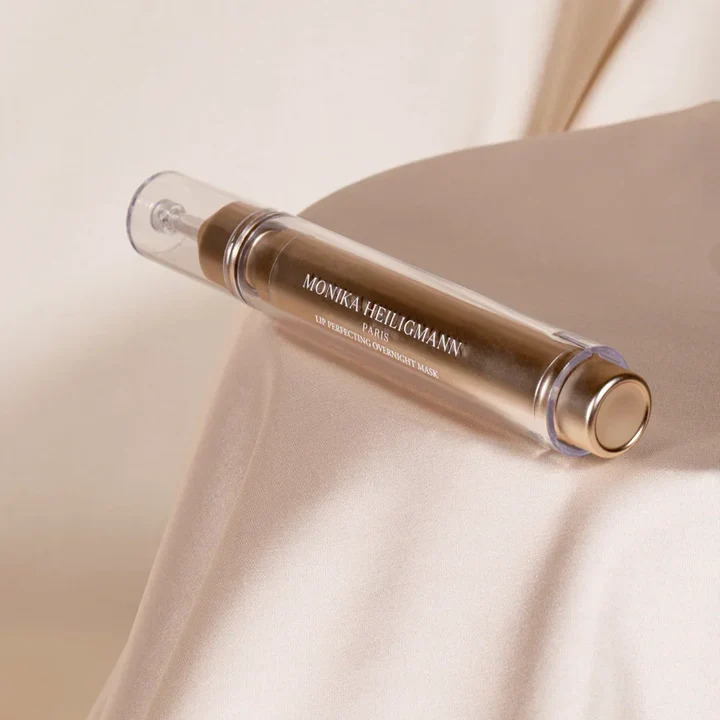 Lydia Esthetics Embassy A Monika Heiligmann Paris lip serum in a clear tube rests on a smooth, beige fabric surface. New York