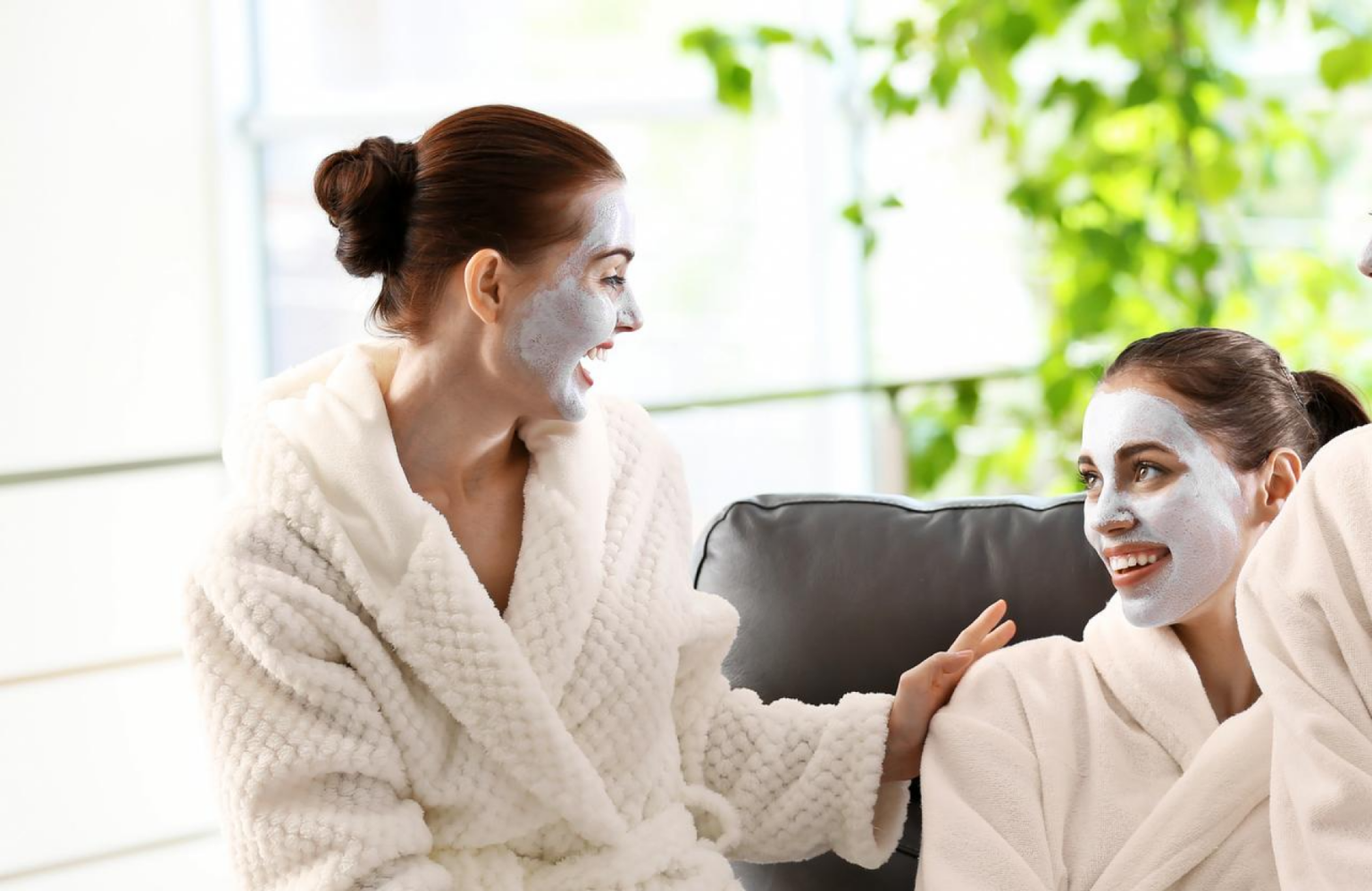 Lydia Esthetics Embassy Two women in white robes with face masks smile and chat while sitting indoors, surrounded by natural light and greenery. New York