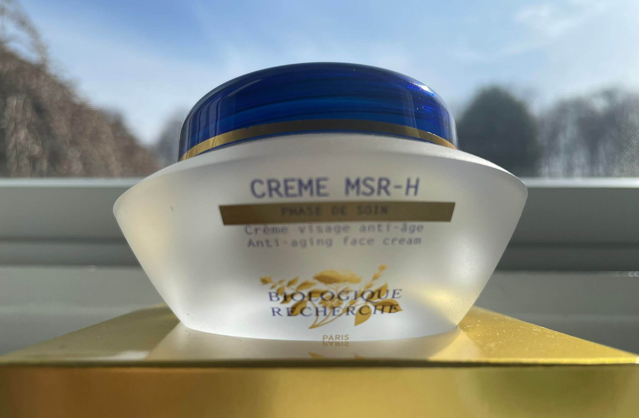 Lydia Esthetics Embassy A jar of Biologique Recherche Creme MSR-H anti-aging face cream sits on a yellow surface in front of a window with trees and sky in the background. New York